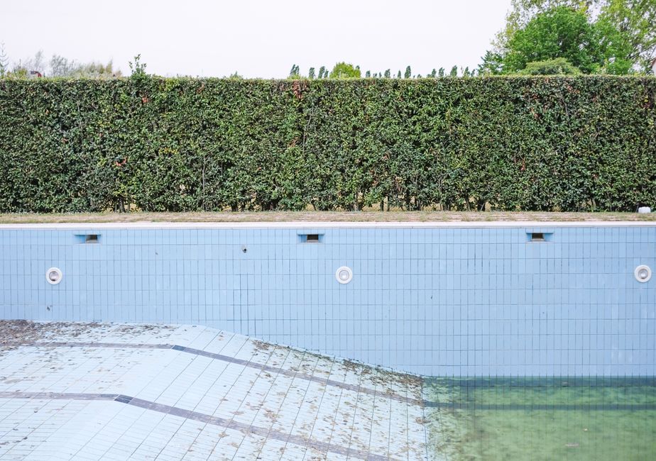 A close-up view of an empty outdoor swimming pool with light blue tiled walls, showing signs of neglect such as dirt, algae, and debris at the bottom and along the edges. The pool's surface is partial